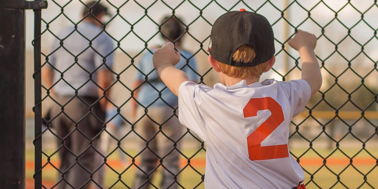 Child behind a fence staring out at a baseball field