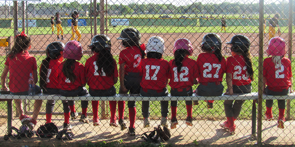 Little League Softball team sitting on the bench in the dugout looking at the field